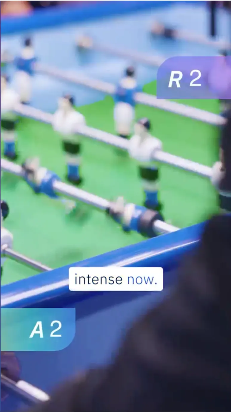 Close-up of a foosball table with live AI score overlays reading R2 and A2 and the caption “intense now.”